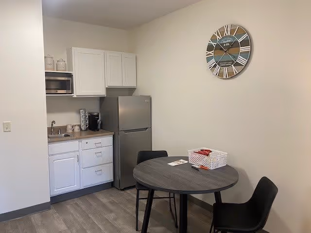 Small kitchen area with white cabinets, a stainless steel refrigerator, a microwave, a coffee maker, and a sink. Next to the kitchen is a round table with two black chairs. A decorative wall clock hangs on the beige wall above the table.