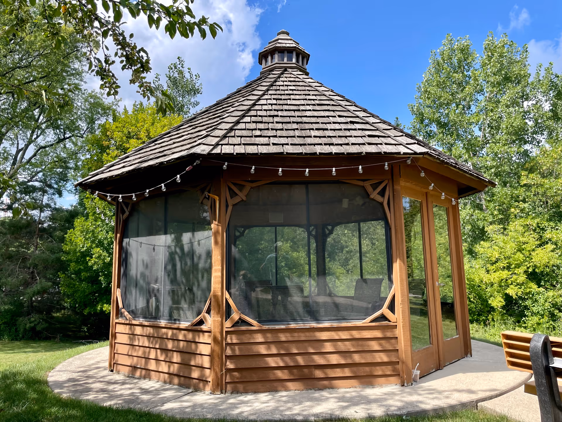 A wooden gazebo with a shingled roof and screened windows, surrounded by green trees and grass under a blue sky with some clouds. There are string lights hanging around the roof edge and a bench partially visible on the right side.