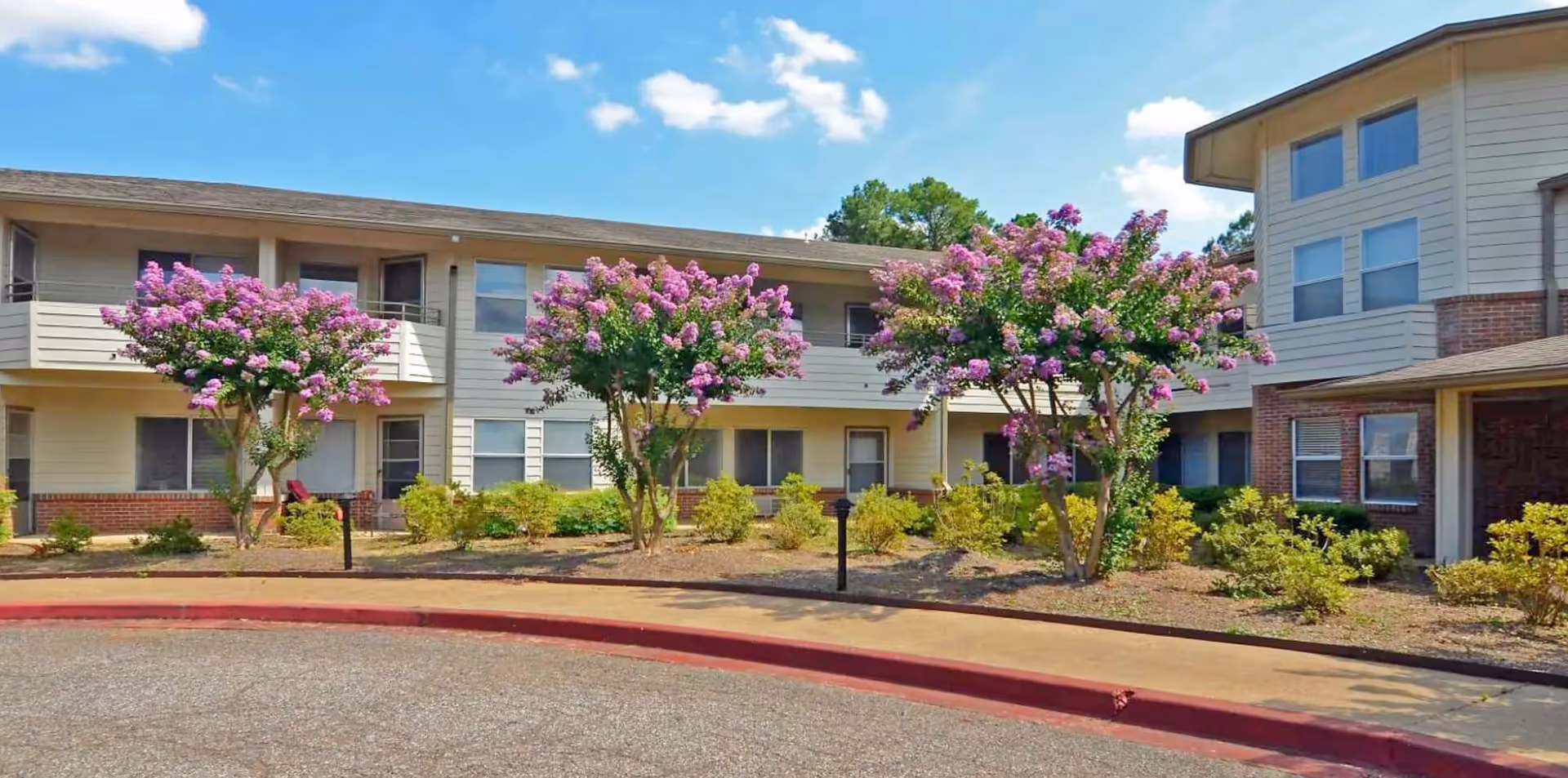 Front of a two-story senior living building with pink flowering trees and landscaped beds along a curved driveway.