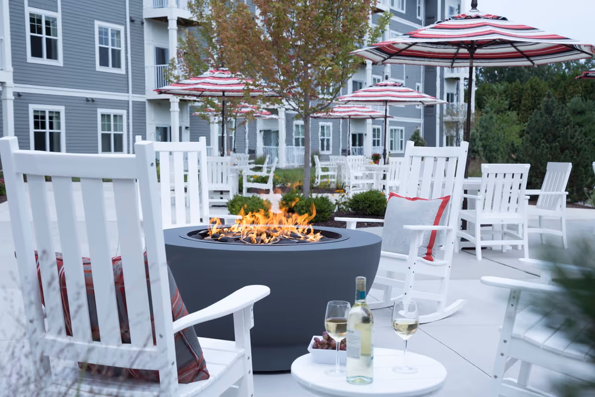 Outdoor patio area at a senior living facility with white rocking chairs, a round fire pit with flames, striped umbrellas, and a small table holding a bottle of white wine, two glasses of wine, and a bowl of grapes. The background shows a multi-story building with gray siding and white trim.