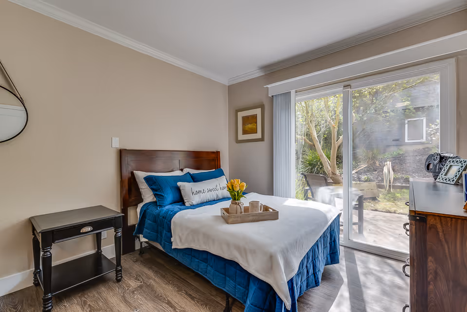 A bright bedroom with a neatly made bed dressed in blue bedding, a tray of flowers on the bed, a nightstand and dresser, and sliding glass doors opening to a patio.