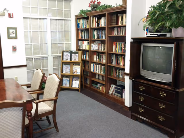 A cozy interior room with a wooden table and upholstered chairs on the left, a tall bookshelf filled with books against the wall, and a wooden cabinet with a television on top on the right. The room has carpeted flooring and a glass-paneled door in the background.