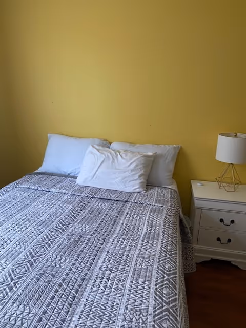 A neatly made bed with a patterned gray and white quilt, two white pillows, and one smaller white pillow against a yellow wall. To the right of the bed is a white nightstand with two drawers and a modern lamp with a white shade and gold geometric base.