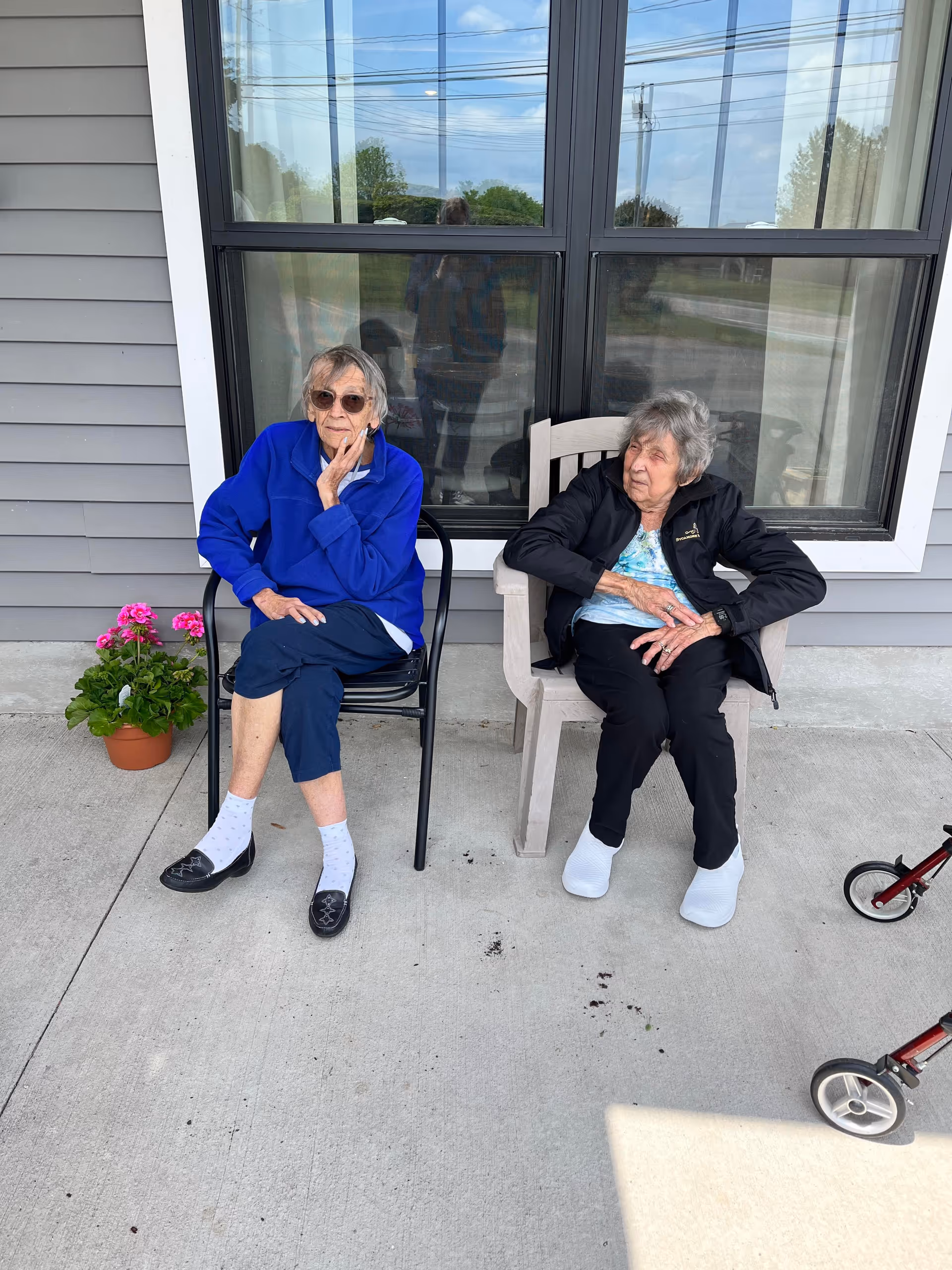 Two older women sitting on chairs on a concrete porch in front of a building window, with a potted flower and a walker nearby.