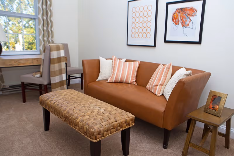 A cozy living room area featuring a brown leather sofa with four decorative pillows in white and orange stripes. In front of the sofa is a woven rectangular ottoman with dark wooden legs. To the right of the sofa is a small wooden side table with a framed photo. In the background, there is a window with patterned curtains and a wooden desk with a chair. Two framed abstract art pieces hang on the wall above the sofa.
