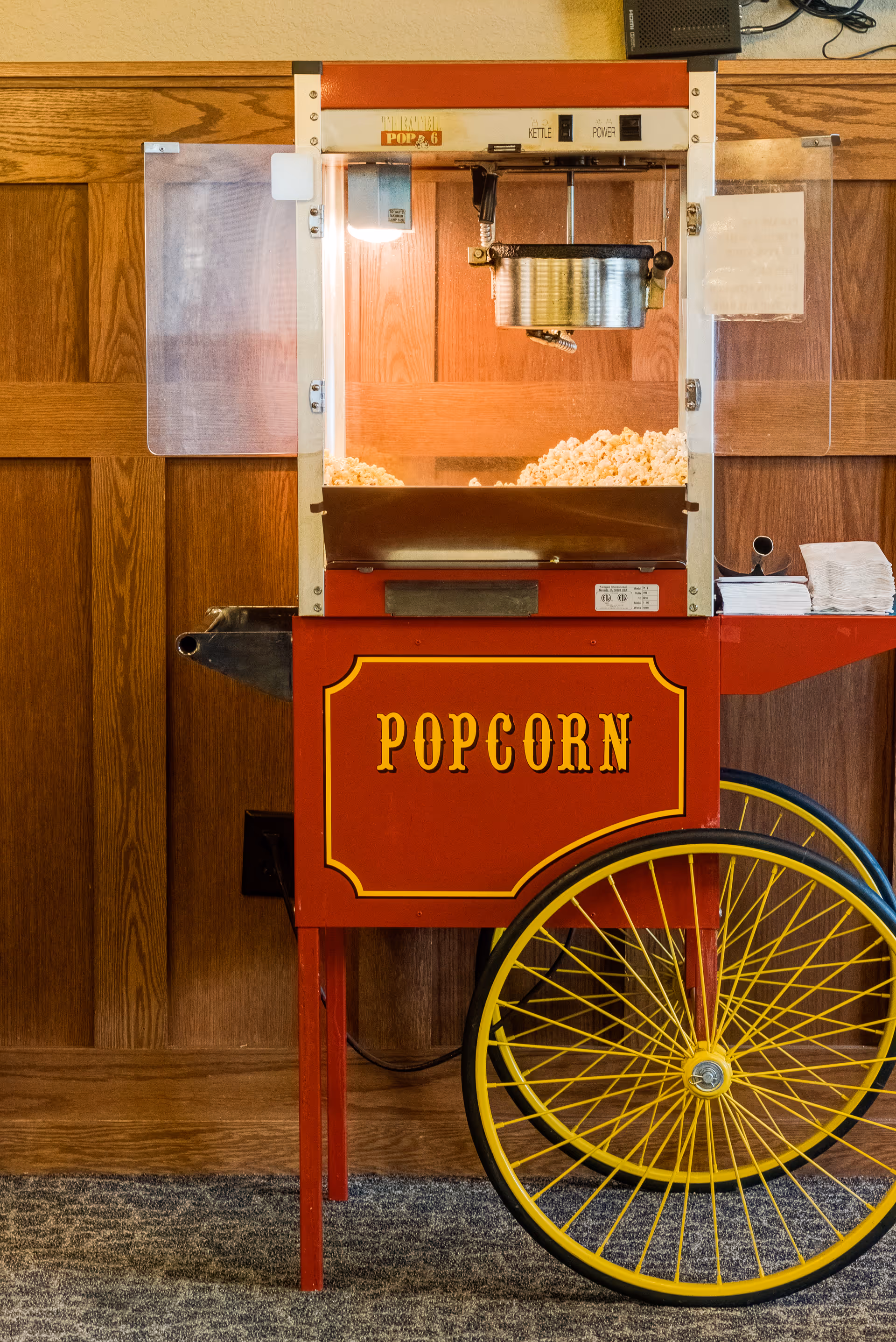 A vintage-style red popcorn machine with yellow wheels, filled with freshly popped popcorn, set against a wooden paneled wall with a carpeted floor.
