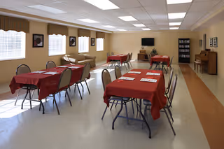 A spacious room with several rectangular tables covered in red tablecloths, each surrounded by chairs. The room has beige walls with windows on one side letting in natural light. There is a beige couch along the wall, a television mounted on the far wall, a bookshelf, and a piano in the corner.