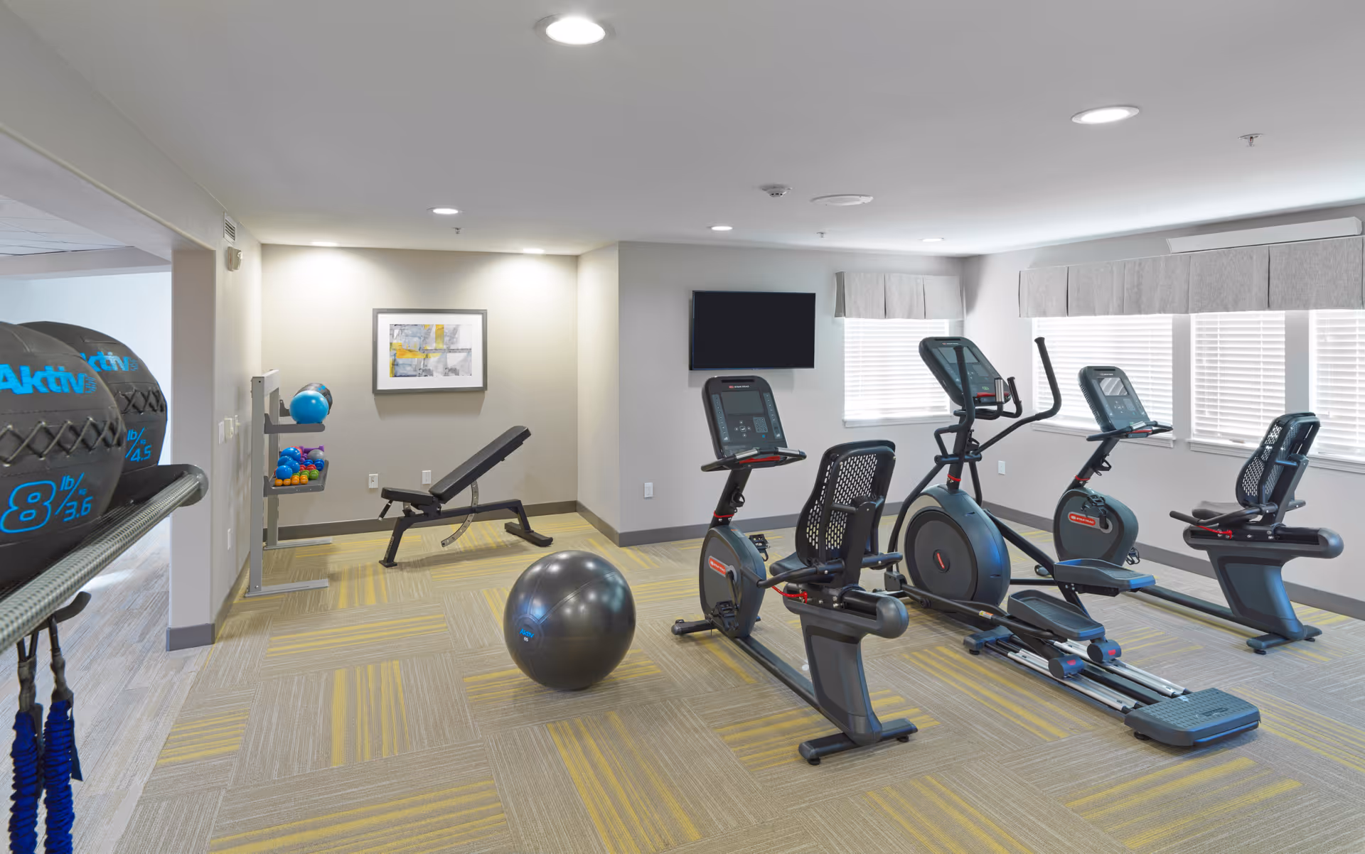 A fitness room in a senior living facility with exercise equipment including stationary bikes, an elliptical machine, a workout bench, medicine balls, and a stability ball. The room has carpeted flooring with a yellow and gray pattern, neutral-colored walls, recessed ceiling lights, and windows with blinds and valances. A flat-screen TV is mounted on the wall.