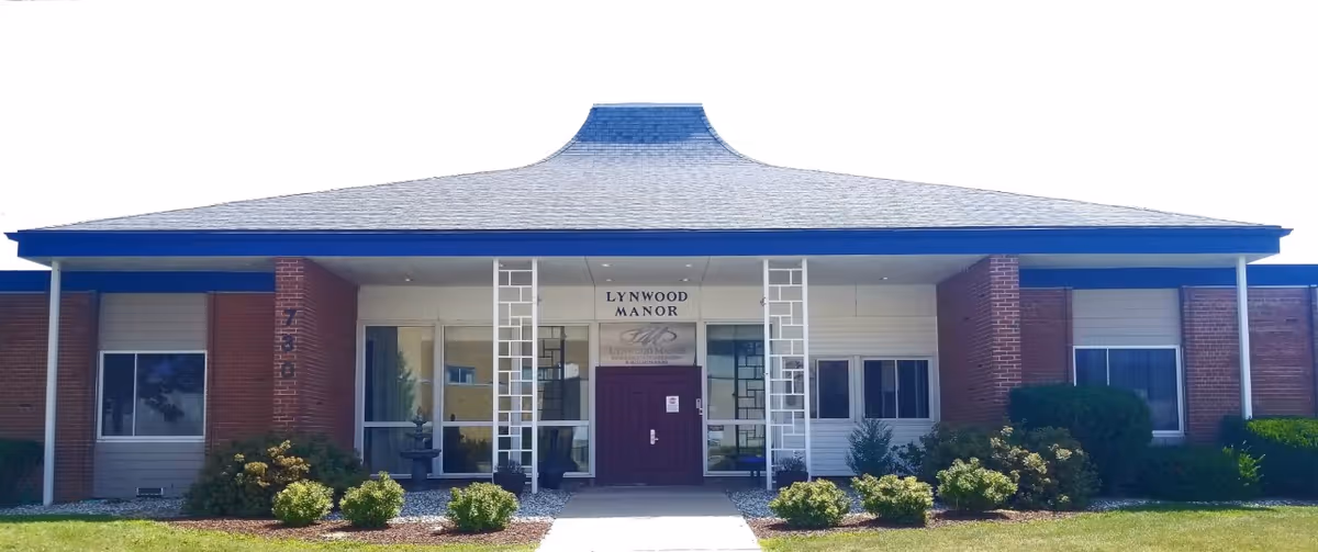 Front exterior view of Lynwood Manor building with a large roof, a central entrance with double doors, windows on either side, and landscaped bushes in front.