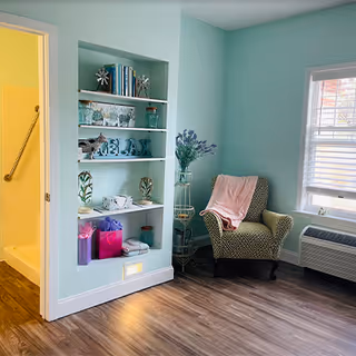 A cozy corner of a room painted in light teal with a patterned armchair draped with a pink blanket next to a window with white blinds. There is a built-in white shelving unit with decorative items including books, glassware, and a sign that says RELAX. The floor is wood laminate, and a bathroom with a yellow wall and a grab bar is visible through an open door.