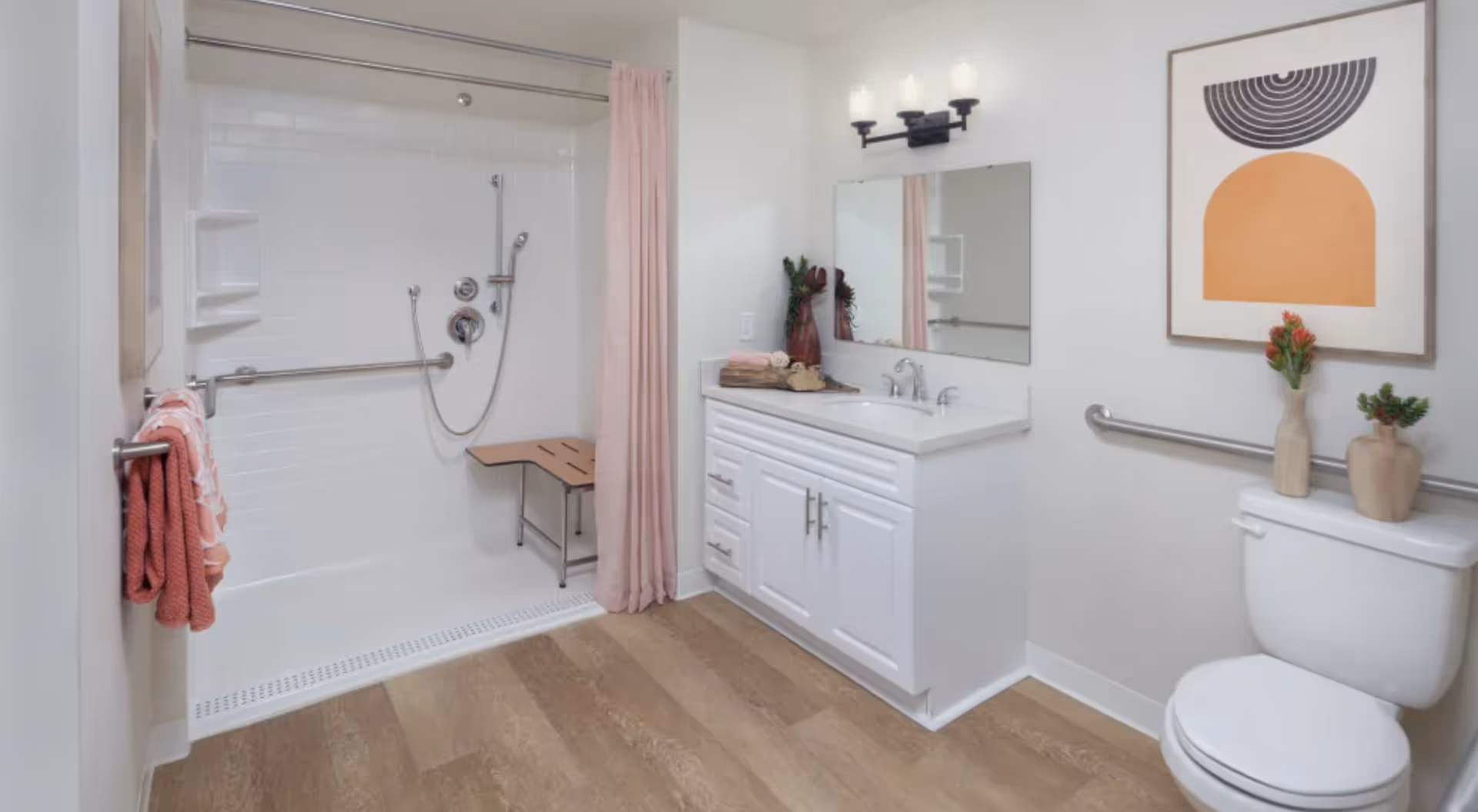 A modern bathroom with a walk-in shower featuring a foldable wooden seat and a handheld showerhead. The shower area has white tiled walls and a pink shower curtain. There is a white vanity with a sink, a large mirror, and a three-light fixture above it. On the right side, there is a toilet with two decorative vases on the tank and a piece of abstract wall art above it. The bathroom has wood-look flooring and grab bars installed near the shower and toilet for accessibility.