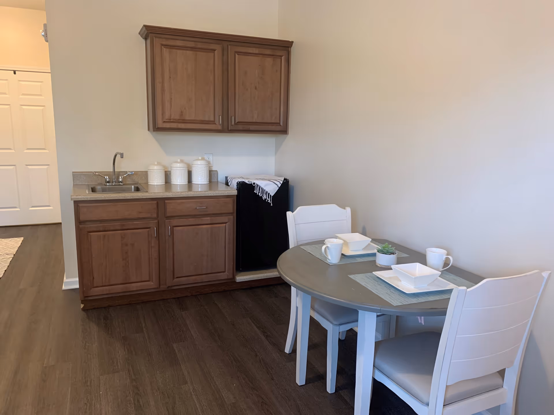 A small kitchenette area with wooden cabinets, a countertop with a sink, three white canisters, and a mini refrigerator. Next to the kitchenette is a round dining table set for two with white chairs, placemats, white mugs, square bowls, and a small potted plant. The floor is dark wood, and the walls are light-colored.