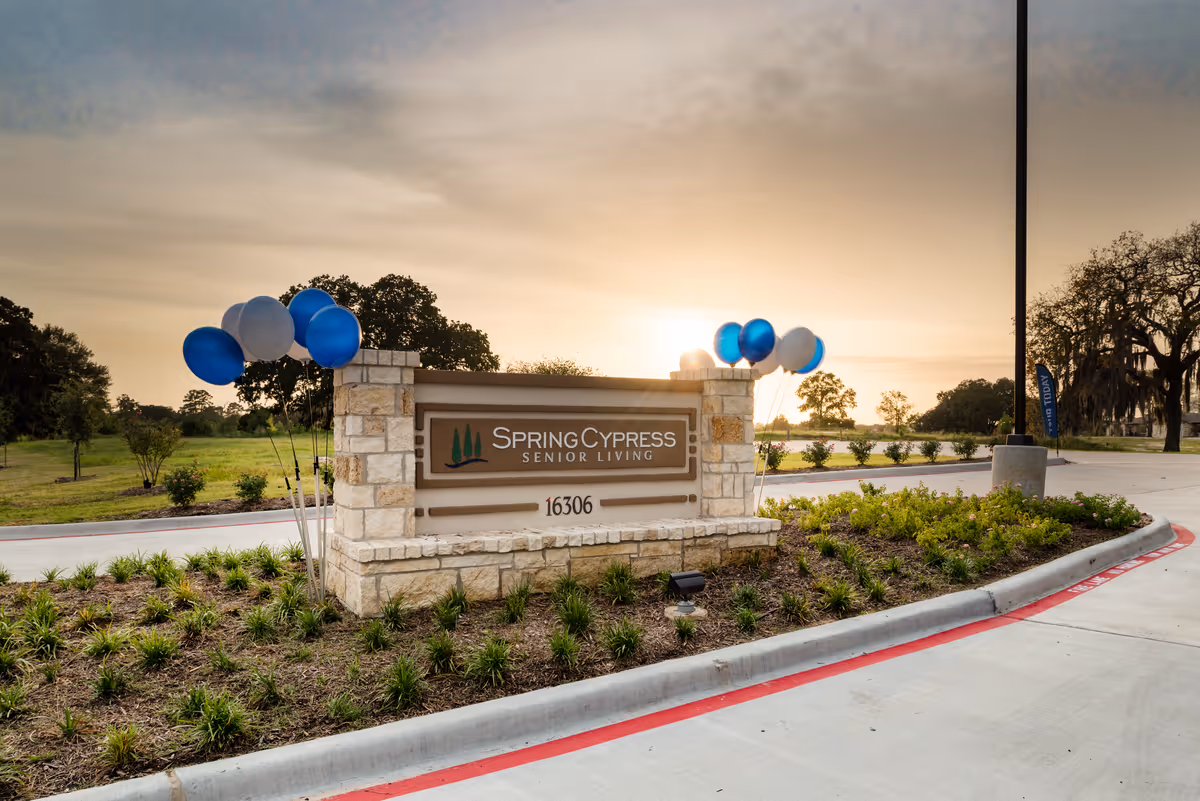 Stone sign for Spring Cypress Senior Living with the address 16306, surrounded by landscaping and blue and white balloons, with a sunset sky in the background.