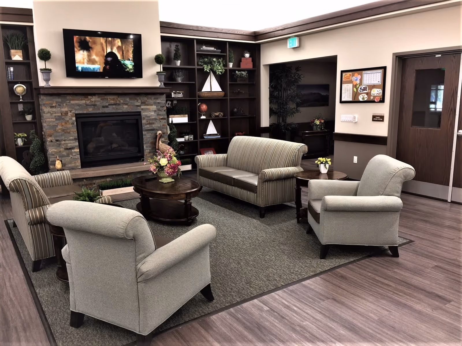 A cozy living room area with a stone fireplace and a mounted flat-screen TV above it. The room features a striped sofa, two matching armchairs, a round wooden coffee table with a floral arrangement, and built-in dark wood shelves decorated with plants and nautical-themed items. The floor is covered with a gray rug and wood-like flooring surrounds the seating area. There is a door and a bulletin board on the wall to the right.