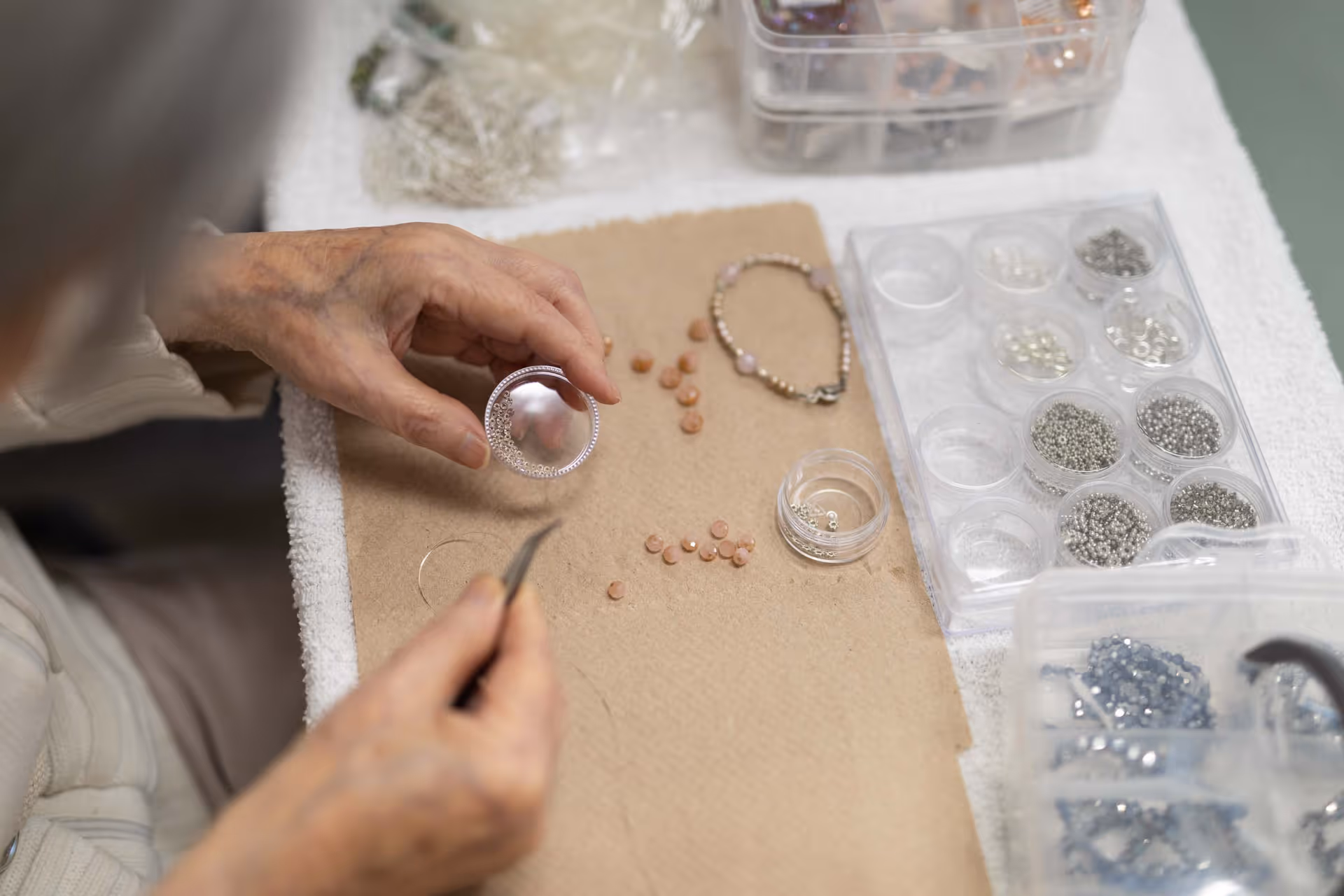 Close-up of an elderly person’s hands working on jewelry making, holding a circular piece with small beads, with various beads and jewelry-making supplies spread out on a table covered with a beige cloth.