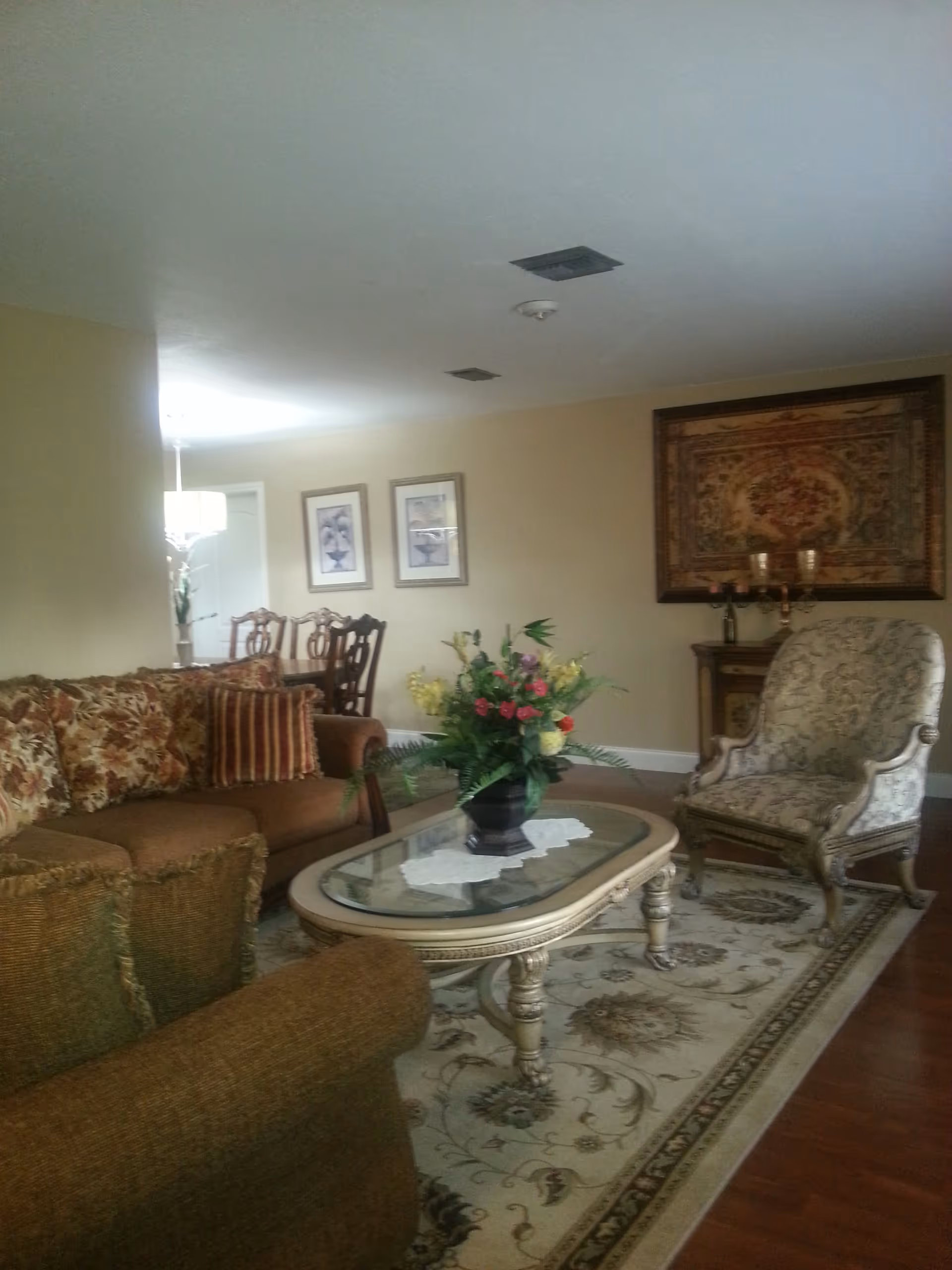 Living room with patterned sofa and armchair around a glass-top coffee table holding a floral centerpiece, with dining chairs and wall art in the background.
