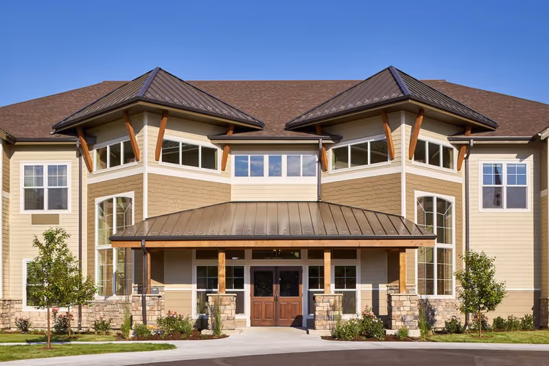 Front exterior of a two-story assisted living building with a covered entrance, large windows, and landscaped grounds.