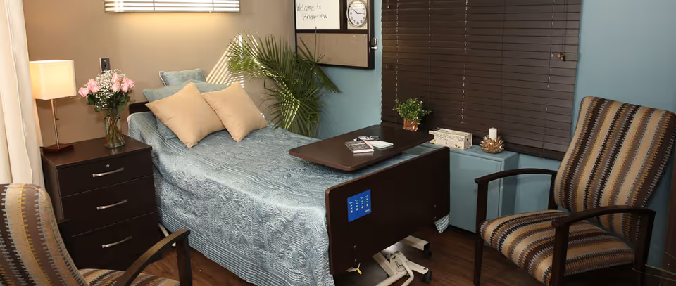 A private resident bedroom with a single hospital-style bed, overbed table, bedside nightstand with lamp and flowers, two armchairs, and window blinds.