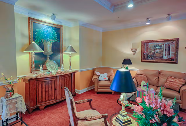 A cozy living room with a red carpet, beige walls, and crown molding. The room features a wooden sideboard with two lamps and decorative items, a framed painting of a vase with flowers above it, a beige armchair with a bunny pillow, a beige sofa with cushions, a glass coffee table with a blue lamp and a floral arrangement, and another framed painting on the wall.
