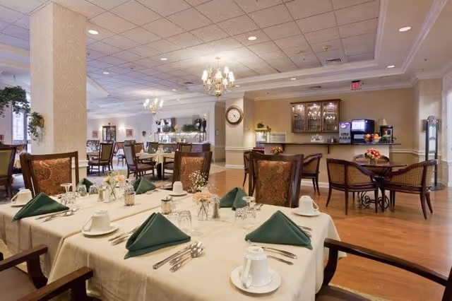 A spacious dining room in a senior living facility with tables set for a meal. The tables are covered with white tablecloths and have green folded napkins, cups, glasses, and silverware neatly arranged. The room features wooden chairs with patterned upholstery, chandeliers hanging from the ceiling, and a beverage station with a coffee machine and fruit on the counter in the background.