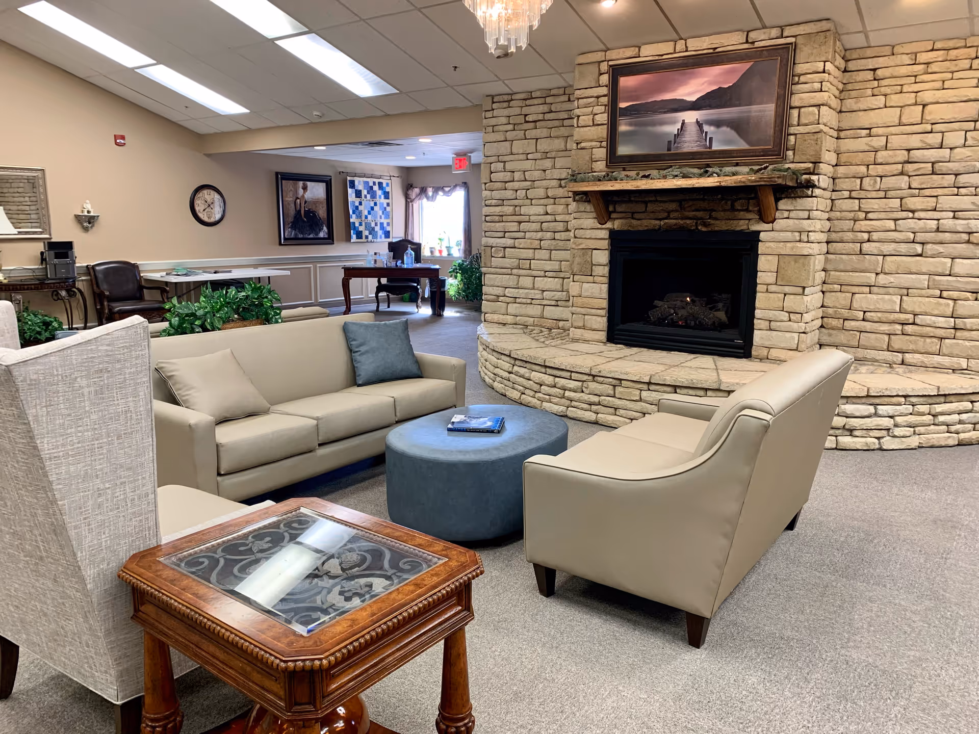 A cozy living room area in an assisted living facility featuring a stone fireplace with a framed picture above it, two beige sofas, a blue ottoman, a wooden side table with a glass top, and various plants and artwork in the background.
