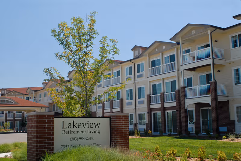 Exterior view of Lakeview Retirement Living building with multiple balconies and large windows under a clear blue sky. In the foreground, there is a brick sign with the facility's name, phone number, and address, surrounded by green grass and a small tree.