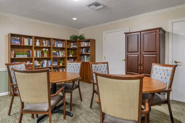 A cozy interior room with a round wooden table surrounded by six chairs with patterned upholstery. Behind the table, there is a large wooden bookshelf filled with books and decorative items. To the right, there is a wooden cabinet and two closed white doors. The room has a carpeted floor and a ceiling with recessed lighting.
