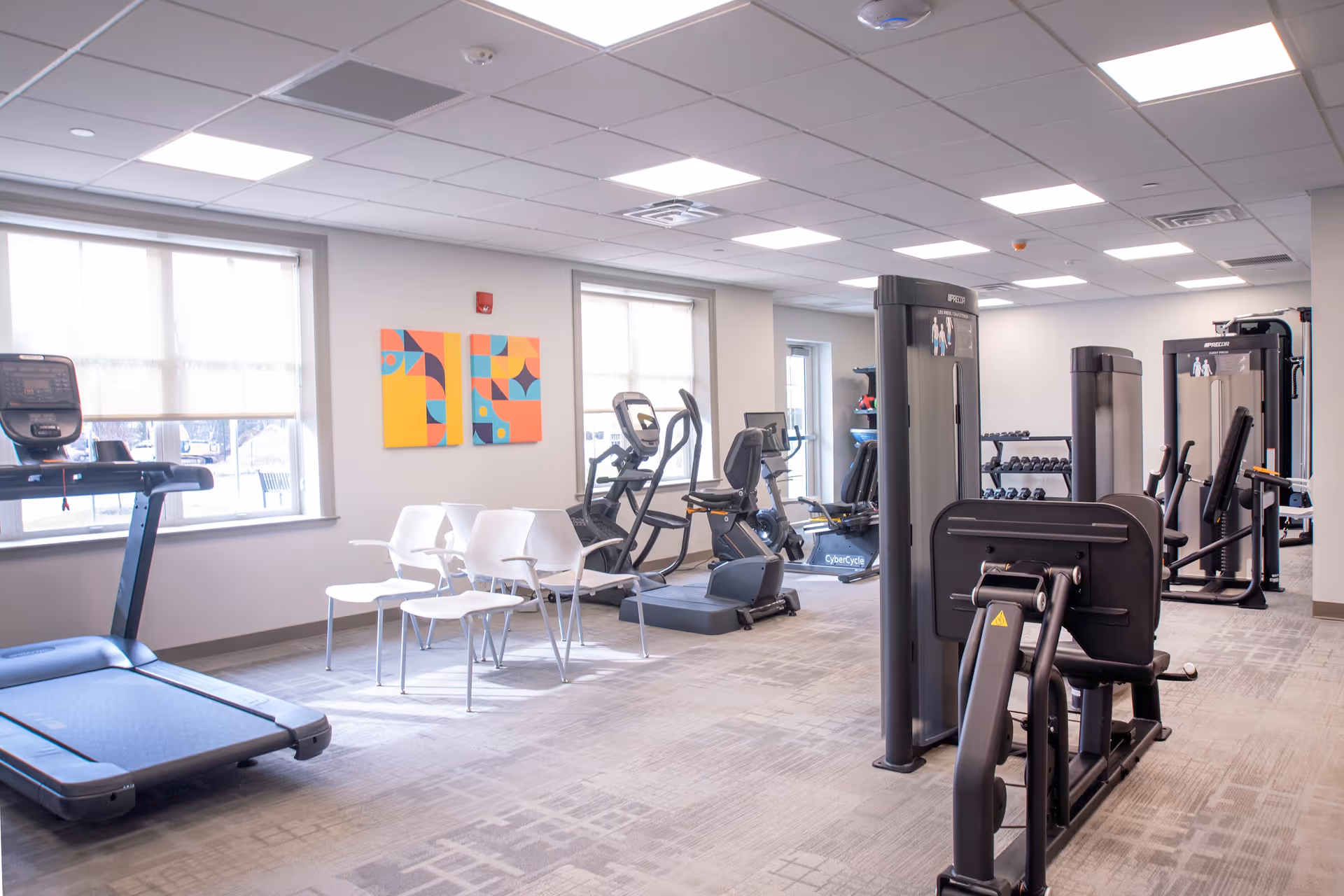 Bright and spacious fitness room with various exercise equipment including treadmills, stationary bikes, weight machines, and free weights. There are large windows with blinds allowing natural light to fill the room, and colorful abstract artwork on the wall. Several white chairs are arranged near the windows.