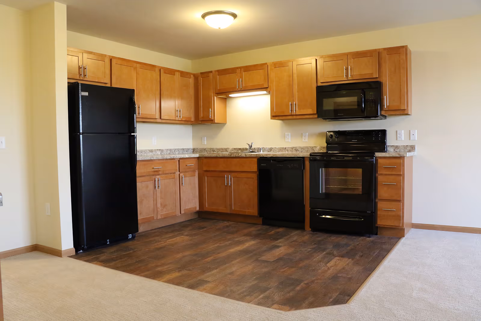 A kitchen area with wooden cabinets, black refrigerator, dishwasher, stove, and microwave. The floor in the kitchen area is wood, transitioning to carpet in the adjacent space. The walls are light-colored and there is a ceiling light fixture.