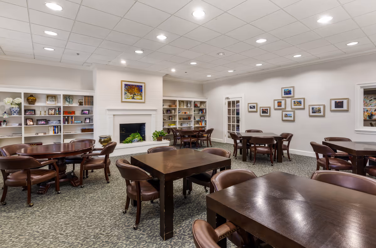 A well-lit common room with multiple dark wooden tables and brown leather chairs on a patterned carpet. The room features a white brick fireplace with a colorful painting above it, built-in white bookshelves filled with books and decorative items, and framed artwork on the walls. The ceiling has recessed lighting, and there is a glass-paneled door on one side.