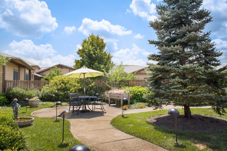 Outdoor garden area with a paved walkway leading to a round patio with a table, chairs, and a large umbrella. The garden is surrounded by green bushes, trees, and a wooden fence under a partly cloudy blue sky.