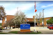 Exterior view of The Embers Retirement Community building with a sign in front, trees, a flagpole with American and Texas flags, and a parked red car under a clear blue sky.
