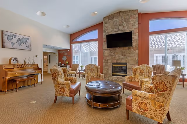 Comfortable senior living common room with patterned armchairs arranged around a round ottoman, a stone fireplace with mounted TV, large windows, and a piano.