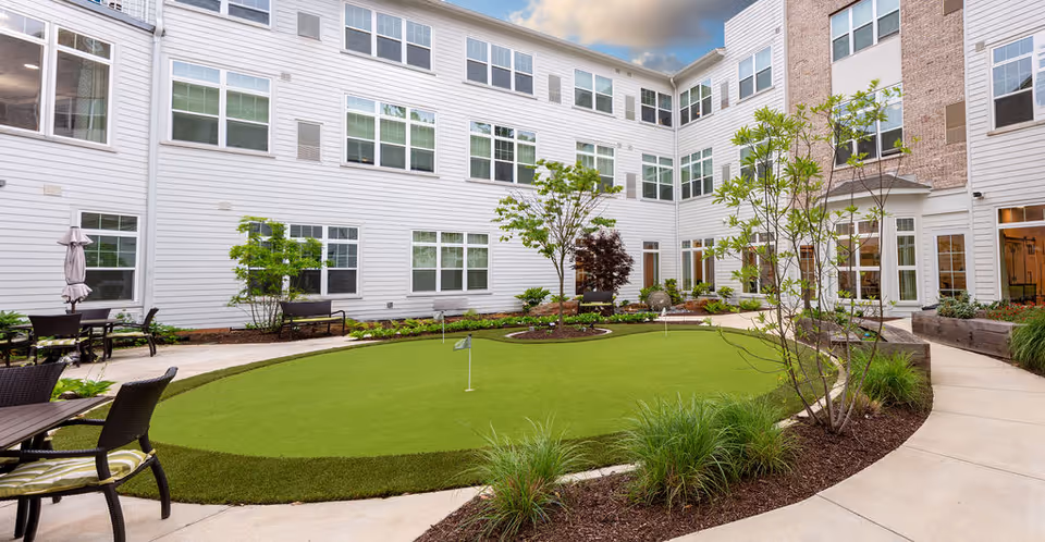 Outdoor courtyard area of The Sheridan at Eastside featuring a putting green surrounded by a curved walkway, patio tables with chairs, small trees, and plants. The courtyard is enclosed by a multi-story building with many windows.