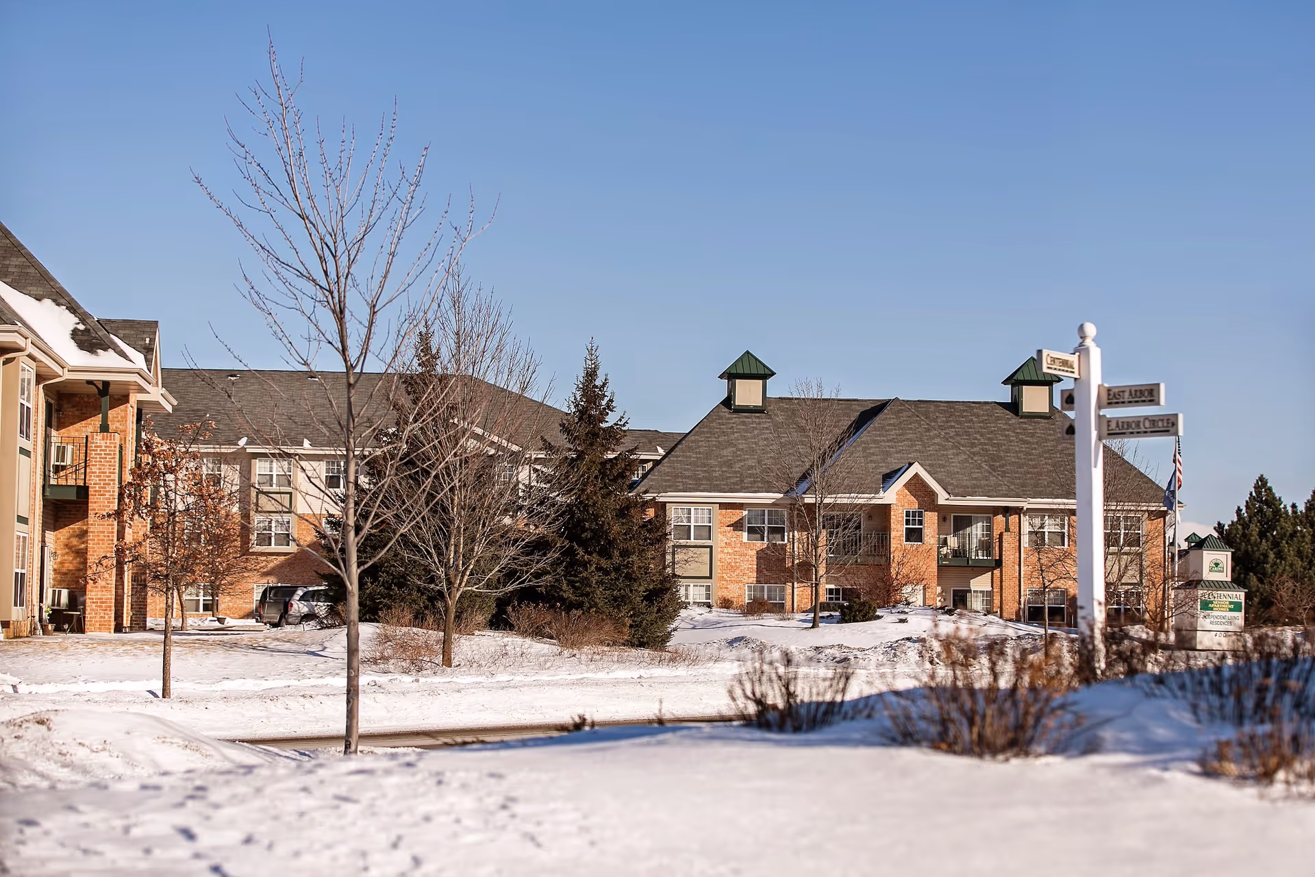 Brick senior apartment building with snow-covered grounds, bare trees, and a signpost in front under a clear blue sky.