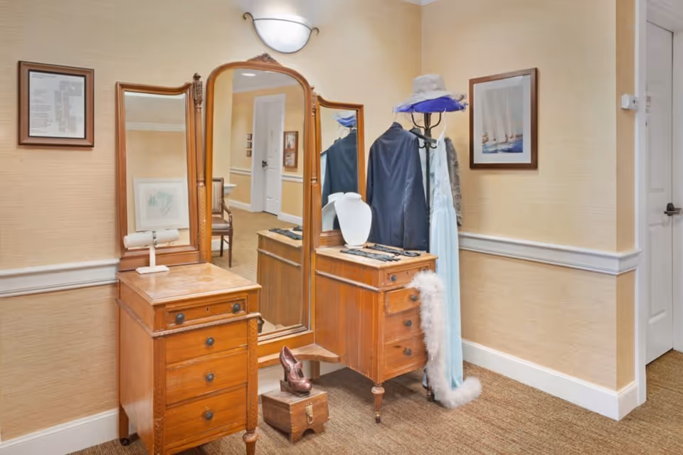 A vintage wooden vanity with three mirrors and multiple drawers is placed against a beige wall with white trim. On the vanity are a white jewelry display bust and some jewelry. Next to the vanity is a coat rack holding a dark jacket, a light blue dress, a white feather boa, and a hat with a purple ribbon. The floor is carpeted, and framed artwork hangs on the walls. A hallway with white doors is visible in the background.