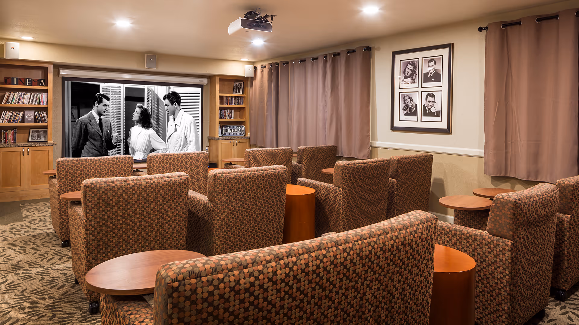 A cozy movie screening room with multiple patterned armchairs and small wooden tables arranged in rows facing a large screen showing a black-and-white film. The room has beige walls, brown curtains, built-in wooden shelves with DVDs, and framed black-and-white portraits on the wall.