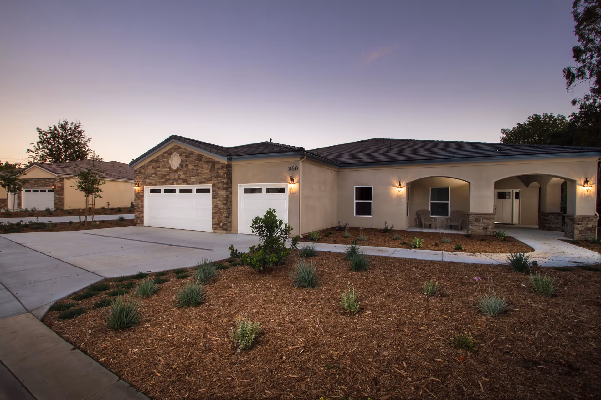 Exterior view of a single-story residential building at dusk with a stone and stucco facade, two white garage doors, a small landscaped front yard with plants and mulch, and a covered porch with two chairs.