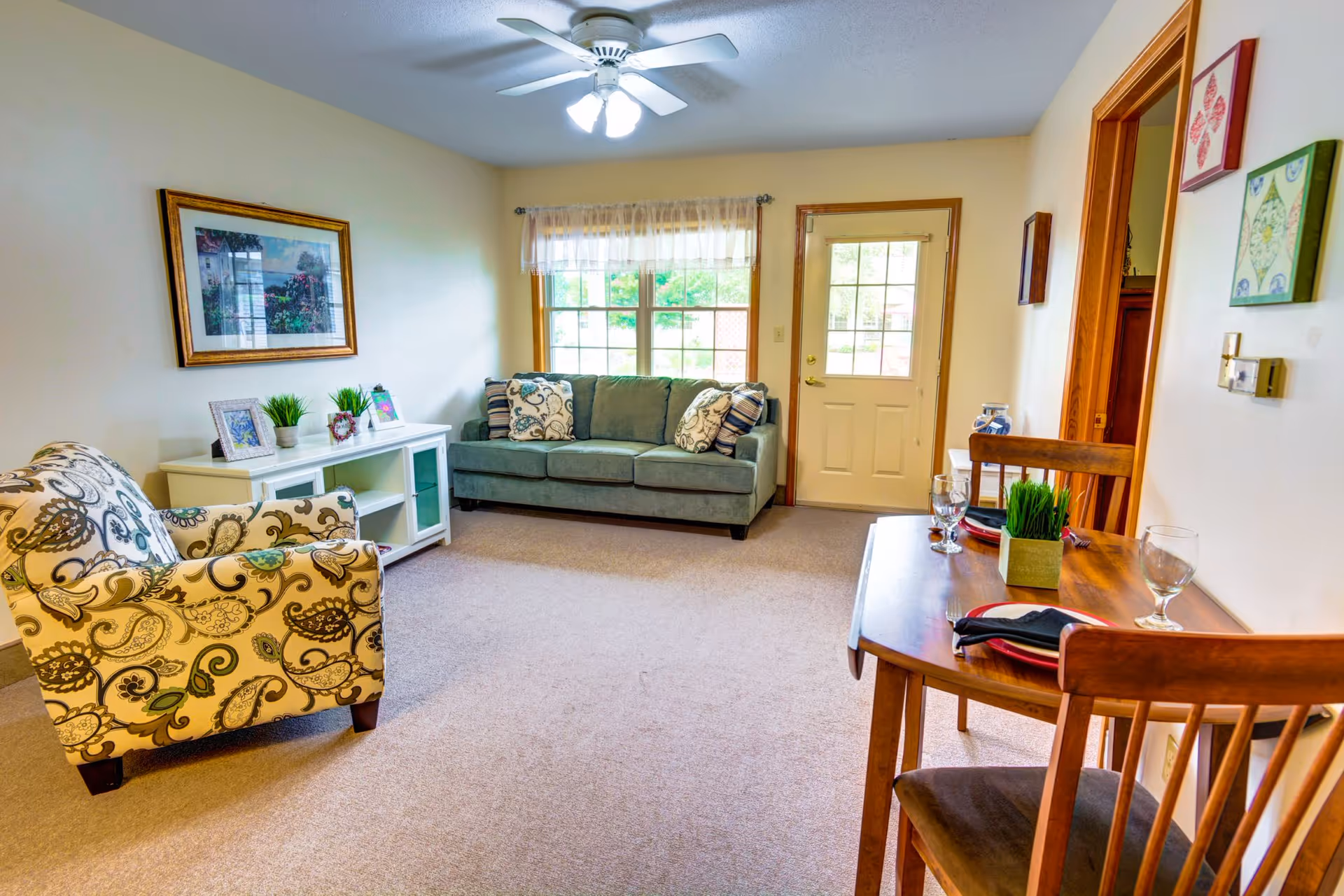 Living room with a patterned armchair, green sofa by a window, a small wooden dining table set, and a ceiling fan.
