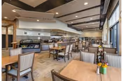 A spacious dining area in a senior living community with multiple wooden tables and cushioned chairs. The room features large windows on one side allowing natural light to fill the space. The ceiling has recessed lighting and wooden beams. In the background, there is a serving counter with food display and kitchen access.