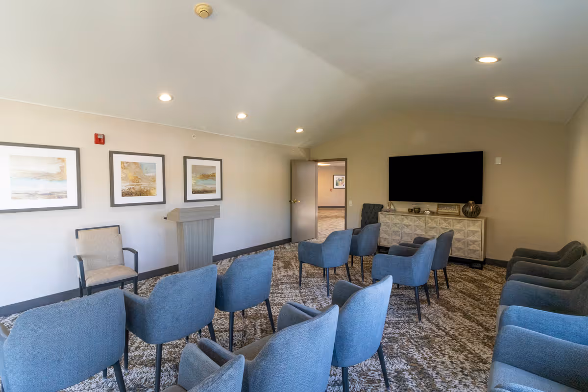 A small conference or meeting room with rows of blue upholstered chairs facing a podium and a large flat-screen TV mounted on the wall. The room has beige walls, a patterned carpet, three framed landscape paintings on one wall, and recessed ceiling lights.
