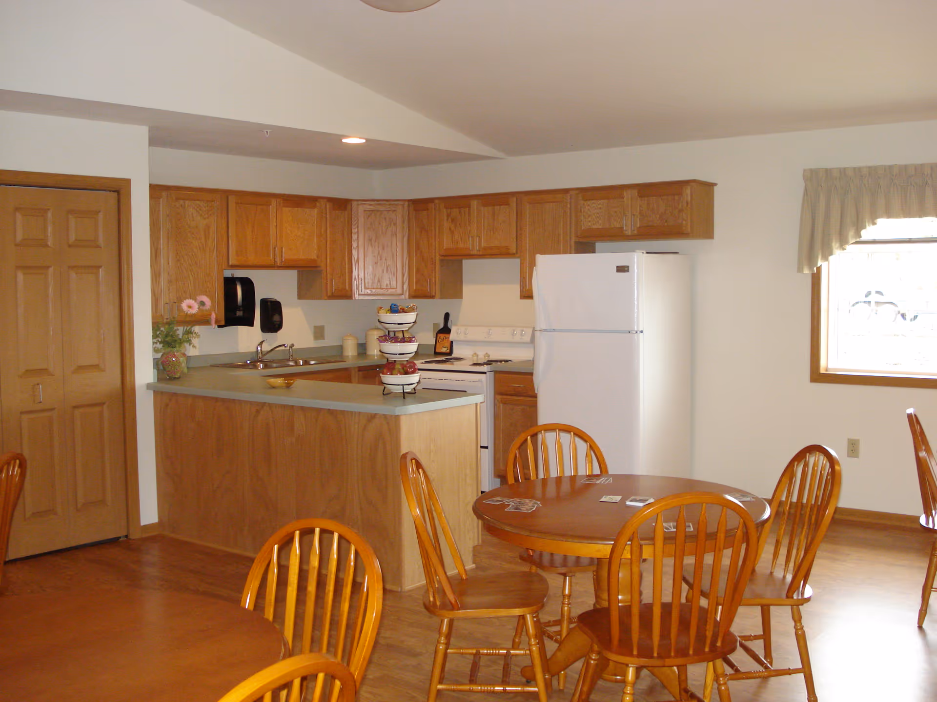 Interior view of a kitchen and dining area in Acorn Hill Apartments featuring wooden cabinets, a white refrigerator, a stove, a sink, and round wooden dining tables with chairs. There is a window with a curtain allowing natural light into the room.