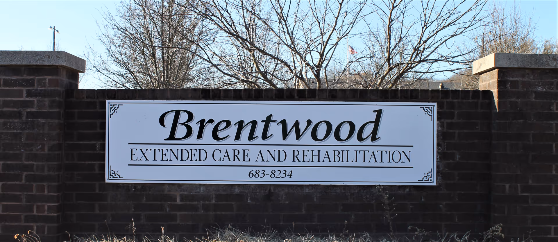 A large white sign with black text mounted on a dark brick wall. The sign reads 'Brentwood Extended Care and Rehabilitation' with a phone number below. Leafless trees and a clear blue sky are visible in the background.