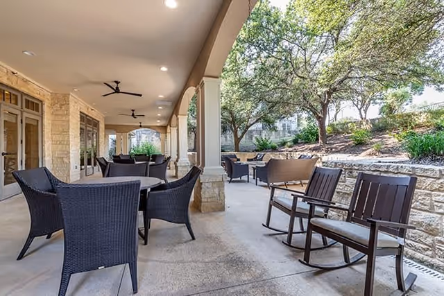 Covered outdoor patio area with multiple seating arrangements including wicker chairs around tables and wooden rocking chairs. Ceiling fans are mounted on the ceiling, and the patio overlooks a landscaped area with trees and stone retaining walls.