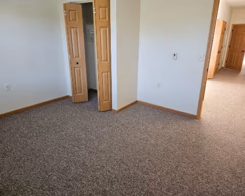Empty room with beige carpet, white walls, wooden trim, and an open closet with wooden bi-fold doors. A hallway with similar flooring and doors is visible in the background.