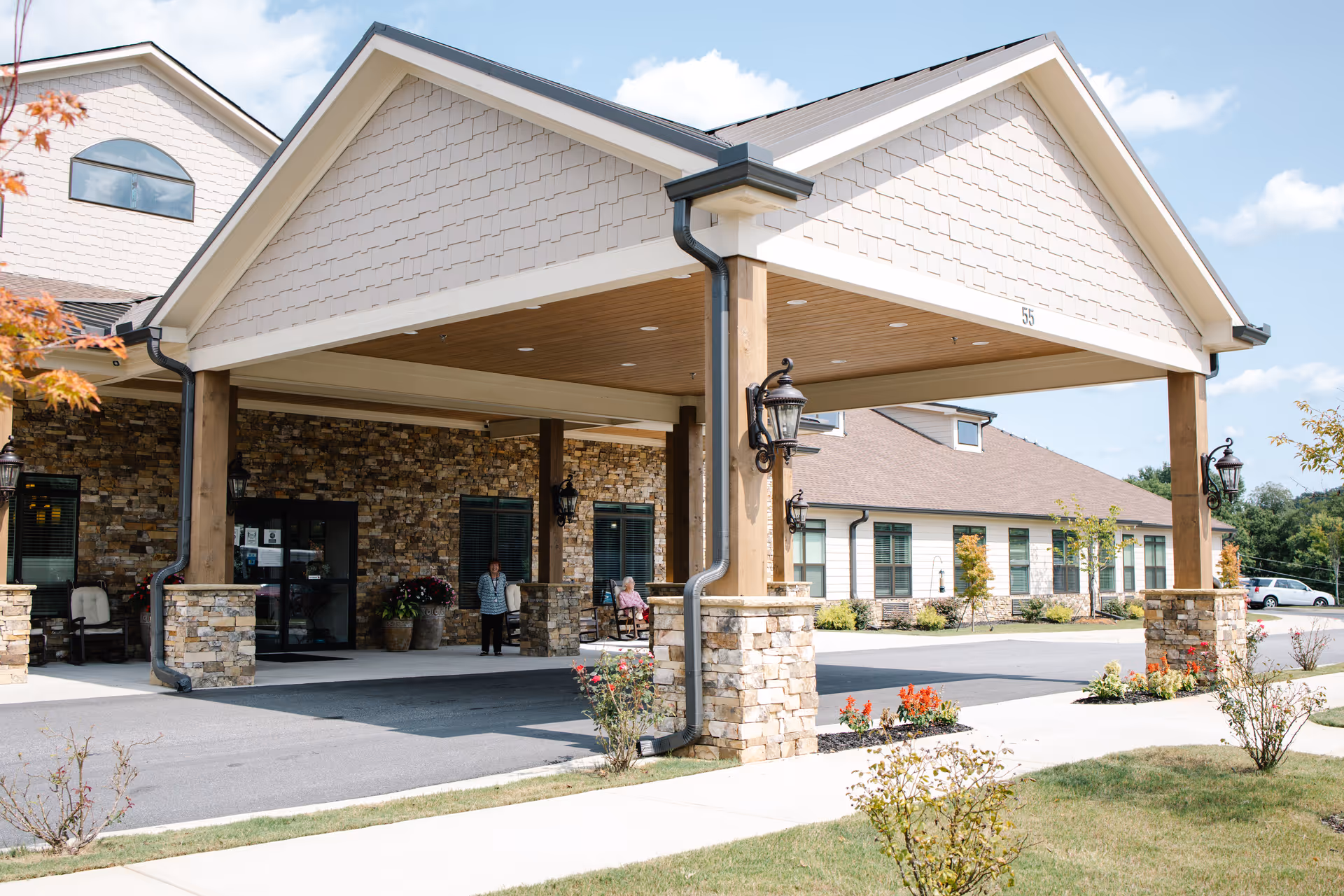 Exterior view of Dahlonega Assisted Living & Memory Care facility entrance with a covered drop-off area supported by wooden pillars and stone bases. Two people are visible near the entrance, and the building has a stone and siding facade with windows and outdoor lighting fixtures. The sky is clear with some clouds.