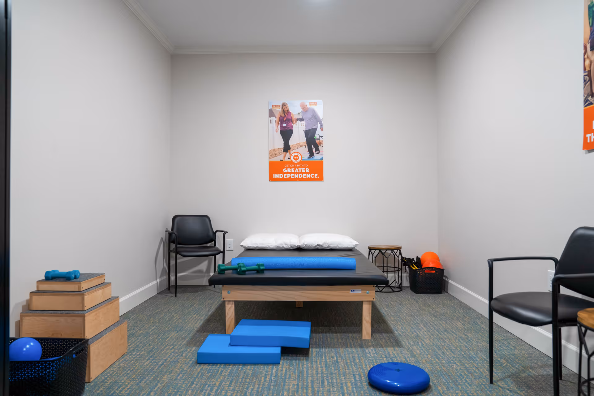 A small therapy or exercise room with a padded treatment table in the center, two black chairs on either side, various exercise equipment including dumbbells, foam rollers, balance pads, and a basket with exercise balls. A motivational poster on the wall reads 'Get on a path to greater independence.'