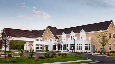 Exterior view of a large two-story brick building with a covered entrance and multiple windows, surrounded by landscaped greenery and a paved driveway under a partly cloudy sky.