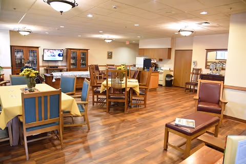 Community dining room with multiple tables covered in yellow tablecloths, wooden chairs, wood flooring, overhead lights, and a TV and cabinets along the back wall.