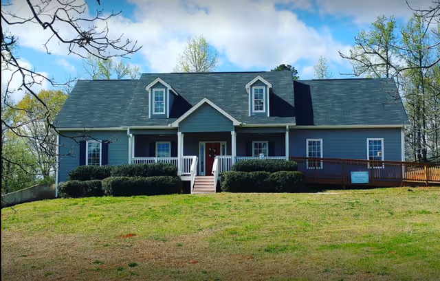 Front exterior view of a single-story blue house with a dark shingled roof, white trim, and a small porch with white railings. There are two dormer windows on the roof and bushes in front of the porch. The house is situated on a grassy lawn with trees in the background under a partly cloudy sky.